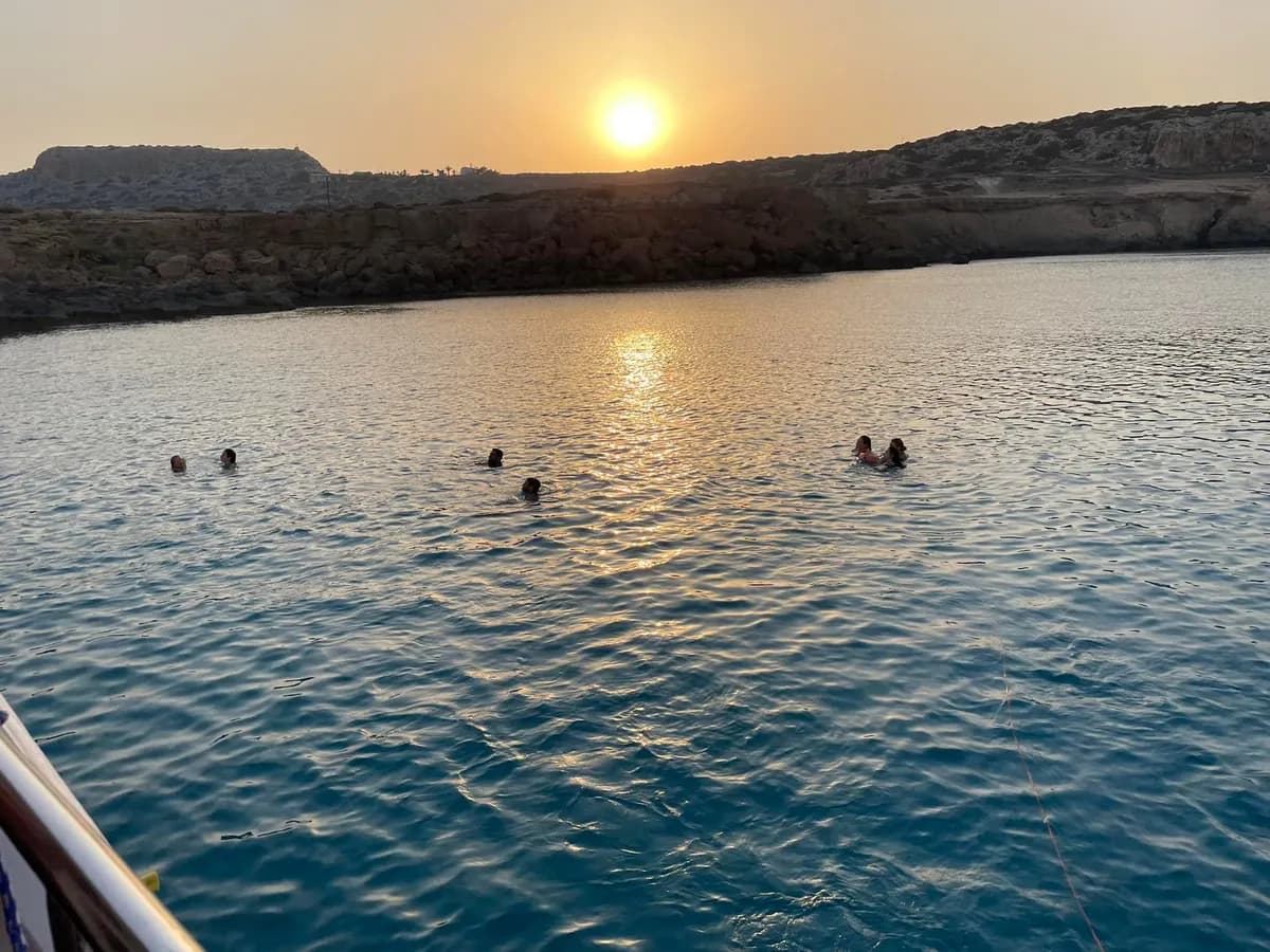 People swimming near a boat as the sun sets beyond the rocky coast